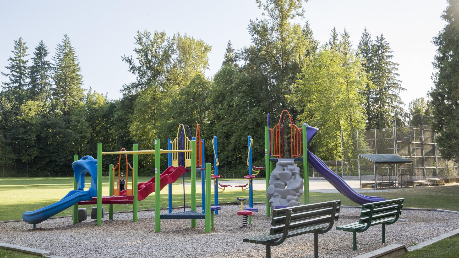 A small colourful playground with two green benches sits overtop a gravel play area with an open field and baseball diamond in back.